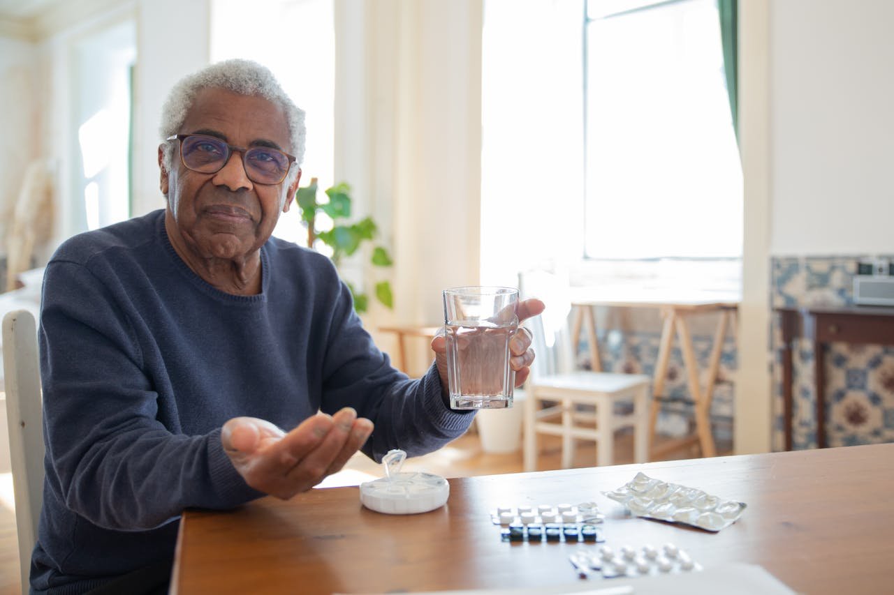 Elderly man sitting at a table at home, holding a glass of water and medication, emphasizing healthcare routine.